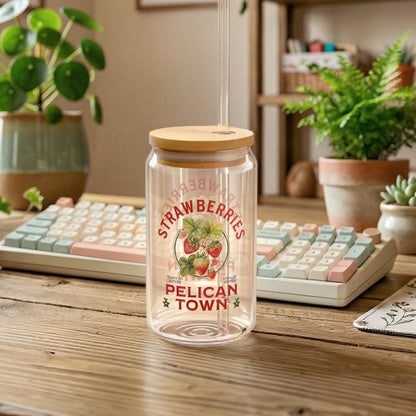 Cozy desk scene featuring a Stardew Valley inspired glass cup with a bamboo lid and straw, decorated with "Strawberries Pelican Town" artwork, set on a wooden table with a pastel mechanical keyboard, houseplants, and warm ambient lighting.
